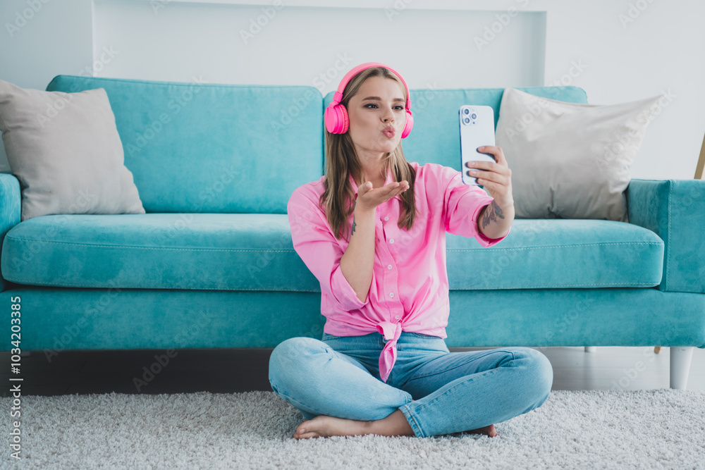 Obraz premium Young woman relaxing at home, blowing a kiss to her phone in vibrant headphones while enjoying leisure time in cozy living room