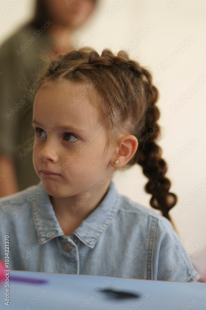 Little beautiful girl in denim shirt sits at table while her hair is being braided. Children's hairstyle concept Little beautiful girl in denim shirt sits at table while her hair is being braided. Children's hairstyle concept
