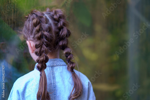 Quadro em tela A little sad girl with two big pigtails stands turned away from the camera near the glass