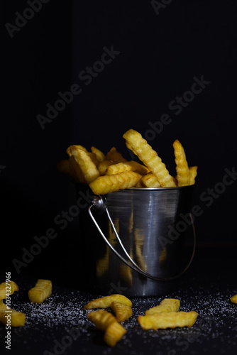 Rippled French fries in a metal bucket on a black background sprinkled with salt