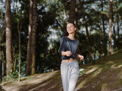 Smiling young woman jogging in the forest, wearing comfortable athletic attire Nature and fitness concept, with green trees and sunlight filtering through branches photo