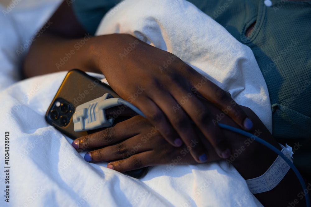 Fototapeta premium Close up shot on African patient's hands with phone in hands and oximeter in hospital ward.