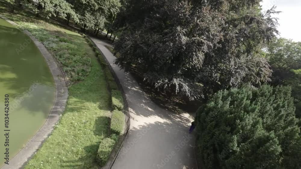 A first-person view FPV flight through a park in Warsaw , Poland. The video starts with a view of a large pond with a fountain in the middle. The camera then flies over a path that winds through the