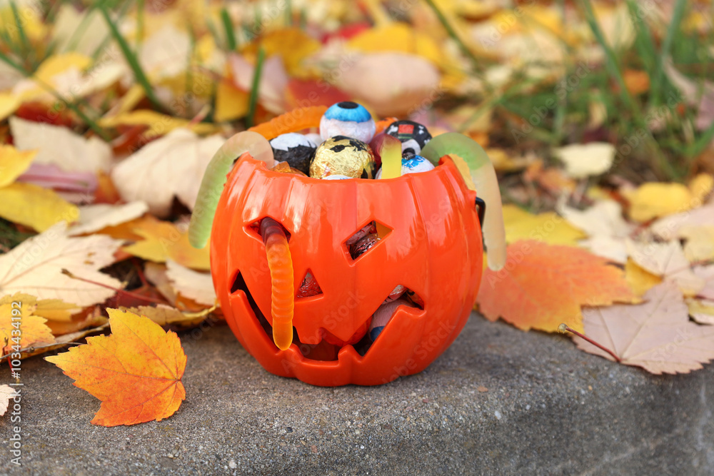 Halloween orange pumpkin jack-o-lantern plastic bucket full of assorted candies and jelly worms, standing on stone curb in autumn park among fallen colorful leaves. Trick or treat concept.