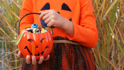 Little girl in orange pumpkin Halloween dress, standing and holding in hands plastic pumpkin with assorted sweets. No face, unrecognizable. Halloween trick-or-treat concept.