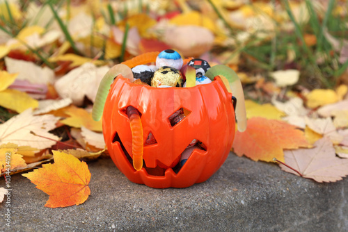 Obraz na płótnie Halloween orange pumpkin jack-o-lantern plastic bucket full of assorted candies and jelly worms, standing on stone curb in autumn park among fallen colorful leaves. Trick or treat concept. w salonie
