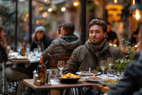 A young man sits at an outdoor table, savoring his dinner in a lively urban restaurant bustling with diners. The warm glow of lights adds to the inviting atmosphere