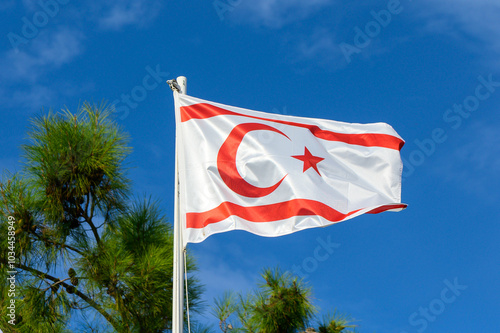 Northern Cyprus flag waving in clear blue sky surrounded by lush green pine trees during bright daylight photo