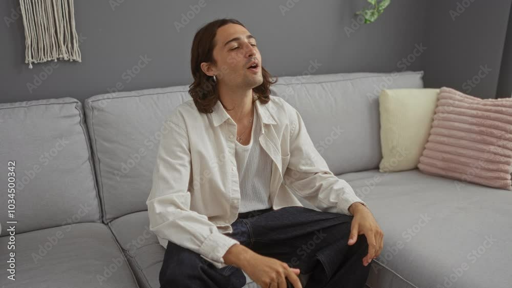 Young man with long hair playing video games on a couch in a living room with a focused and intense expression Young man with long hair playing video games on a couch in a living room with a focused and intense expression