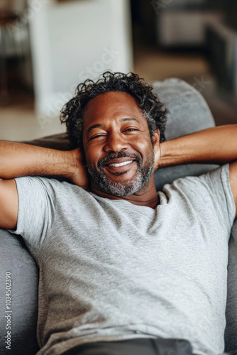 A middle-aged Black man with wavy hair and a well-trimmed beard reclines on a soft couch, smiling widely, his hands behind his head. The serene home ambiance reflects his deep sense of relaxation and