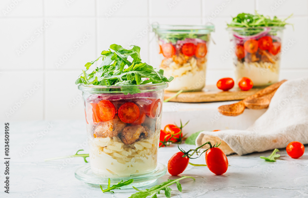 A vibrant layered salad in three glass jars, featuring a fresh mix of dressing, pasta, chicken, cherry tomatoes, red onion, and arugula on a light-colored background. Selective focus. Healthy food A vibrant layered salad in three glass jars, featuring a fresh mix of dressing, pasta, chicken, cherry tomatoes, red onion, and arugula on a light-colored background. Selective focus. Healthy food