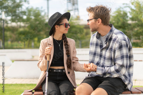 A visually impaired woman and her partner sit together outside, engaging in a warm conversation. They enjoy each other's company, surrounded by a tranquil landscape under a bright sky