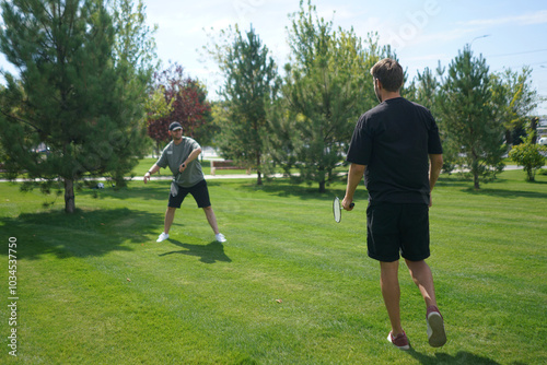 Two individuals engage in a friendly badminton match on a lush green lawn, surrounded by trees under a clear blue sky on a beautiful summer day