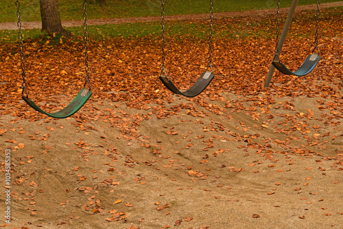 Three empty playground swings with Autumn leaves on ground.