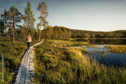 A woman walks along a wooden boardwalk beside the peaceful lake gränjåstjärnen, enjoying the warm glow of summer golden hour with Idre Fjäll ski slopes in the background, Sweden