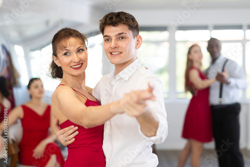 Happy smiling woman dressed in red dress enjoying waltz with male partner during group party in modern latin dance salon. Social dancing concept photo
