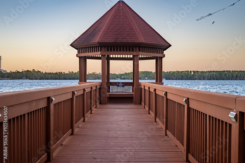 A stunning wooden pier stretches out towards Byron Lake, offering breathtaking views of the serene water and the surrounding lush greenery. The warm tones of the pier, combined with the clear evening 