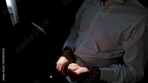 The man sits on a chair around the window and holds two rings in his hand.