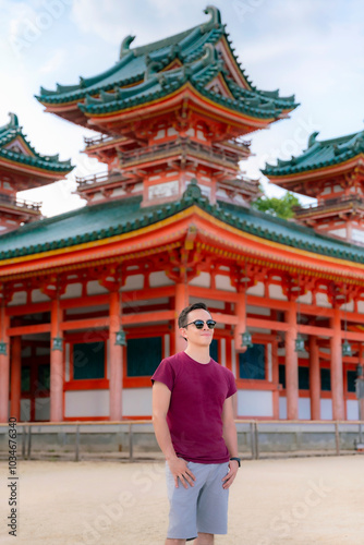 A young happy Latin man poses confidently in front of the iconic Heian Jingū Shrine in Kyoto, Japan. He wears a casual outfit with sunglasses, enjoying his sightseeing experience.