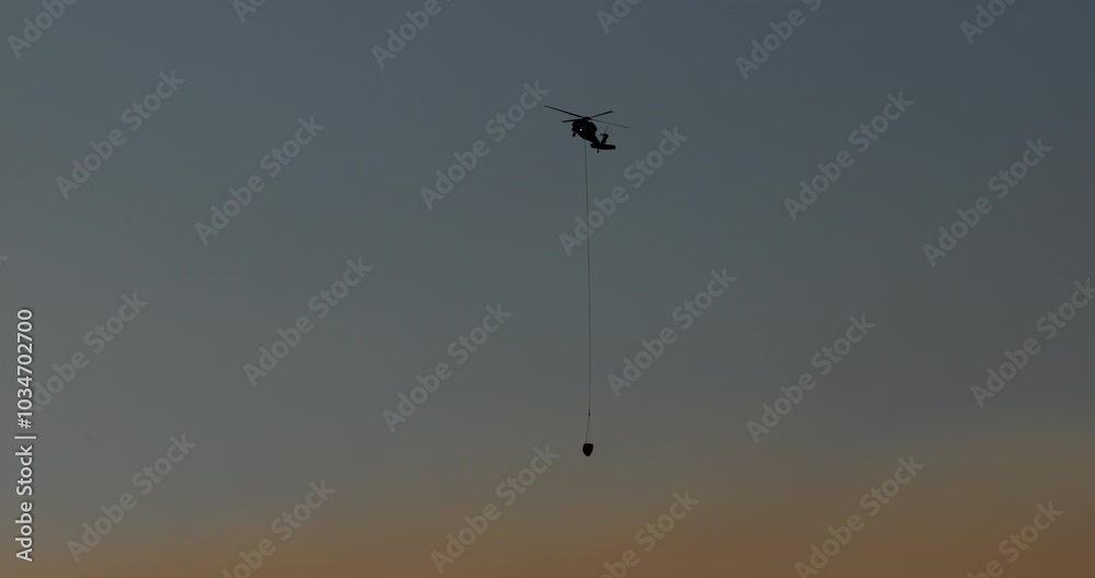 Helicopter with a water bucket . A massive help with local forest fires with a large dense orange cloud of smoke at the bottom.