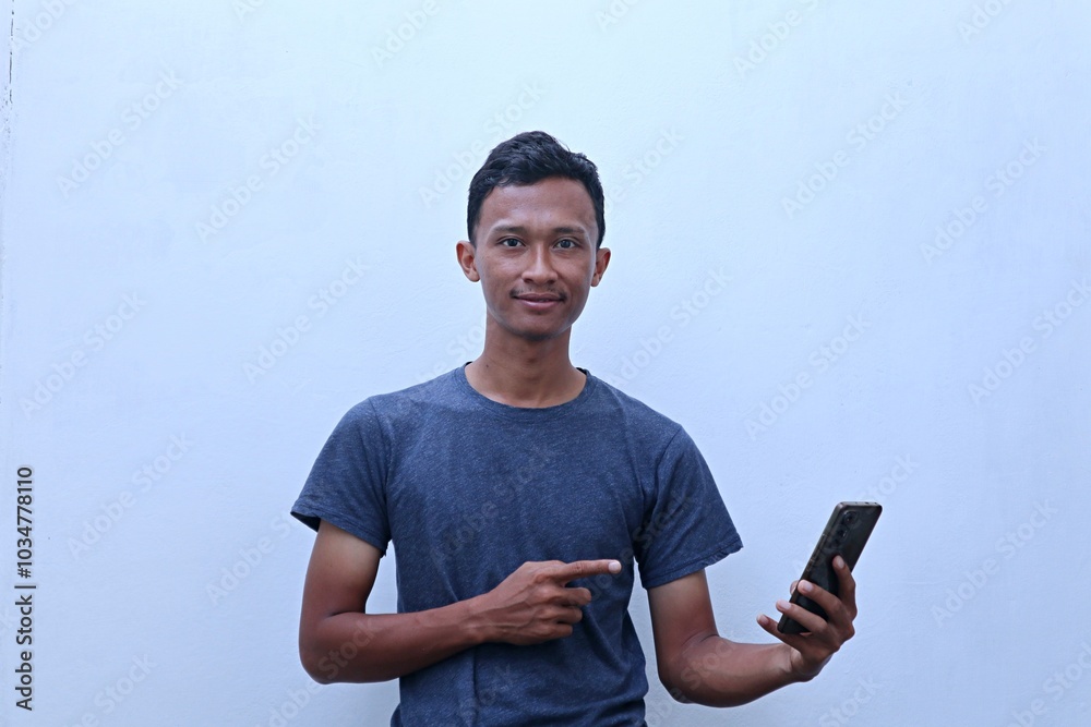 A young Asian man wearing a gray t-shirt holds a smartphone while smiling A young Asian man wearing a gray t-shirt holds a smartphone while smiling