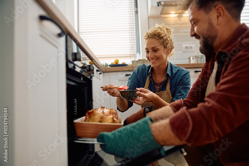 Photos Happy couple basting Thanksgiving turkey with oil while preparing food at home