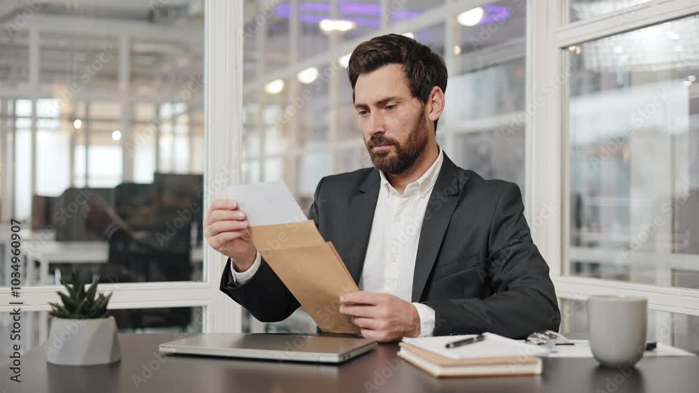 A happy businessman in a suit sits at his desk in the office and opens an envelope with a letter. He is smiling. He is happy with the news. A happy businessman in a suit sits at his desk in the office and opens an envelope with a letter. He is smiling. He is happy with the news.