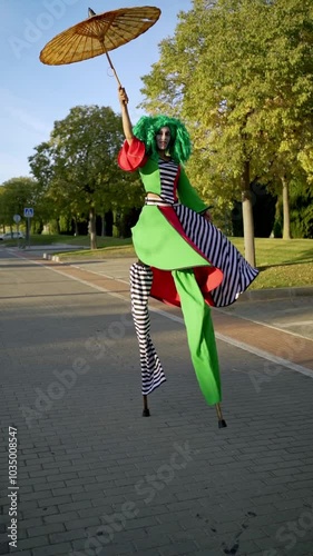 Street performer in a colorful costume gracefully balances a parasol while walking on stilts in a sunny park