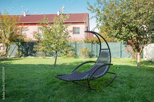 A rocking chair looks like a crescent in the yard of the house on lawn, under the shade of tree in the garden. A relaxing and quiet comfy porch during a sunny day in own backyard of country house photo