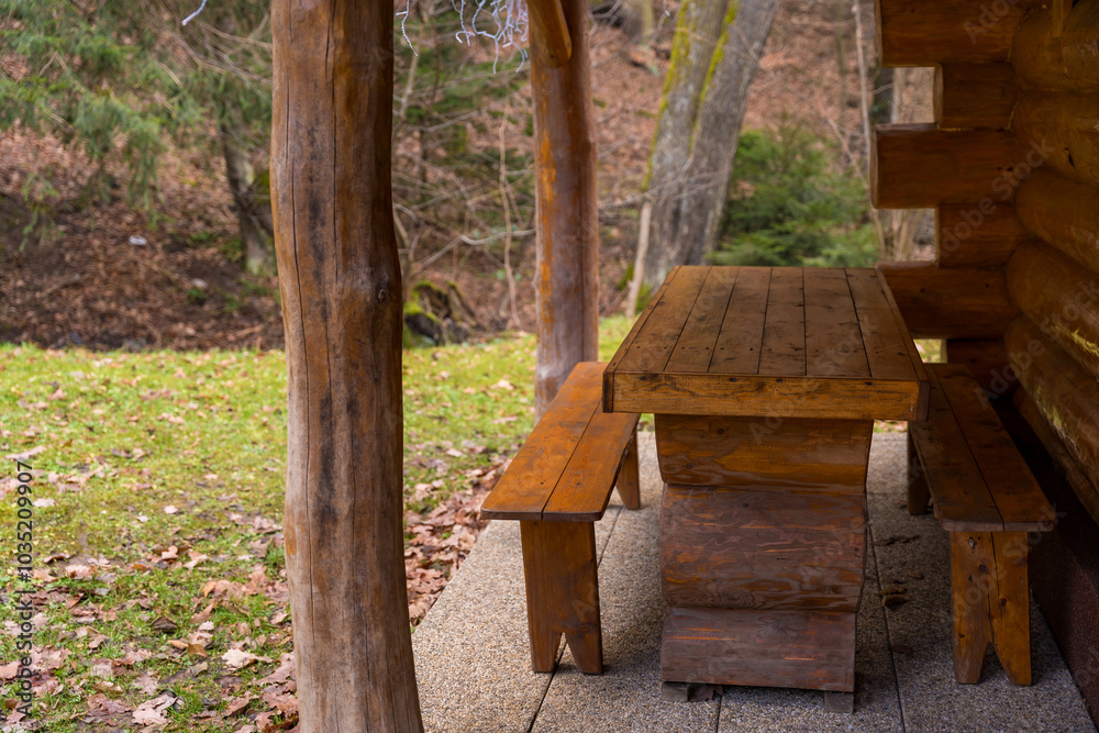 Naklejka premium Rustic wooden table with two benches near a log cabin on a cloudy winter snowless day in a forest.