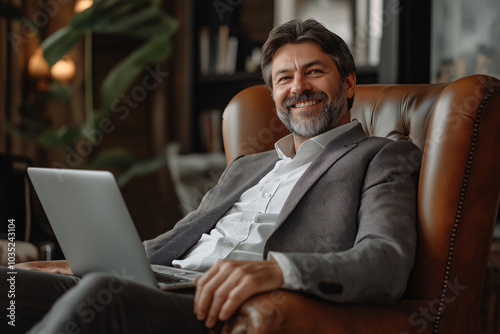 Mature businessman with a beard in a suit looks dignified, with a laptop on his lap, sitting in a brown armchair, smiling charmingly and looking relaxed.