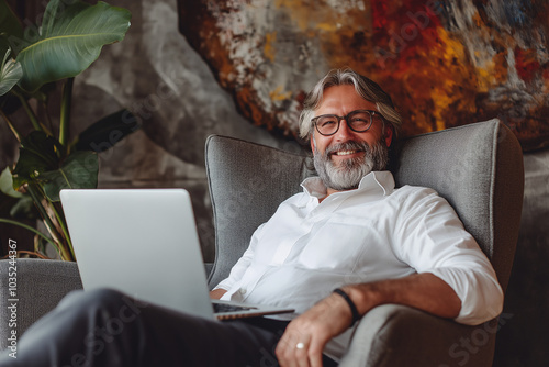 Mature businessman with a beard in a white shirt looks dignified, with a laptop on his lap, sitting in a grey armchair, smiling charmingly and looking relaxed