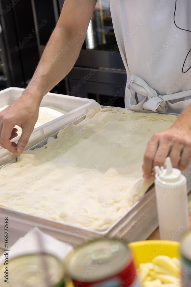 Close-up side view of male cook hands preparing thin rectangular pizza dough on professional kitchen table in pizzeria or italian restaurant. Soft focus. Copy space. Food service business theme. Close-up side view of male cook hands preparing thin rectangular pizza dough on professional kitchen table in pizzeria or italian restaurant. Soft focus. Copy space. Food service business theme.