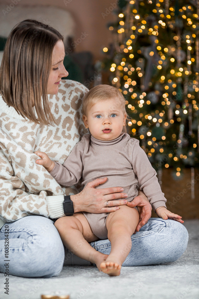 A beautiful little blond boy of 1 year old is sitting with his mother on the background of a Christmas tree. The concept of celebrating Christmas and New Year. A beautiful little blond boy of 1 year old is sitting with his mother on the background of a Christmas tree. The concept of celebrating Christmas and New Year.