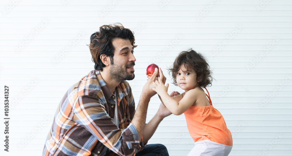 Young bearded man father eating a red apple by hand with his cute daughter. Dad and adorable little girl have fun playing, sitting and standing on floor at home. Happy Caucasian family relaxing Young bearded man father eating a red apple by hand with his cute daughter. Dad and adorable little girl have fun playing, sitting and standing on floor at home. Happy Caucasian family relaxing