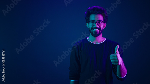 Smiling Indian man amazed Arabian IT guy Muslim male posing pointing index finger on copy space workspace isolated on neon dark blue ultraviolet background in studio showing thumb up coding promotion