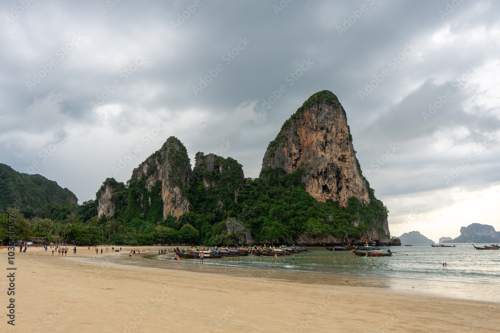 A serene beach with longtail boats and towering cliffs under a cloudy sky. A serene beach with longtail boats and towering cliffs under a cloudy sky.