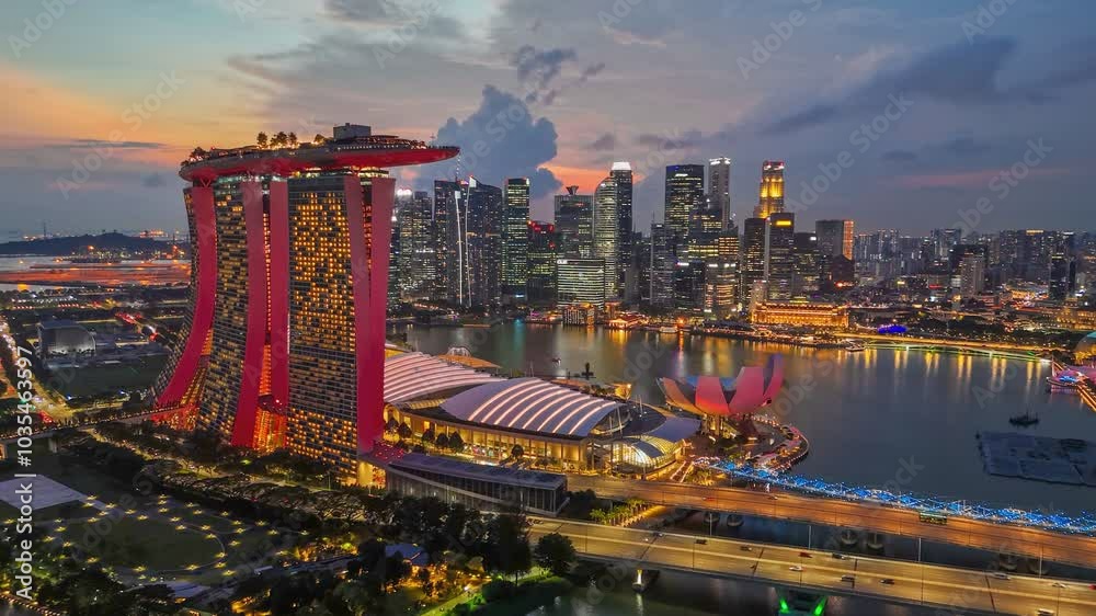 Aerial view of Singapore at dusk. Marina Bay Sands hotel is prominently displayed in foreground. City lit up by streetlights and buildings, reflecting in water of bay. Skyline of Singapore at sunset