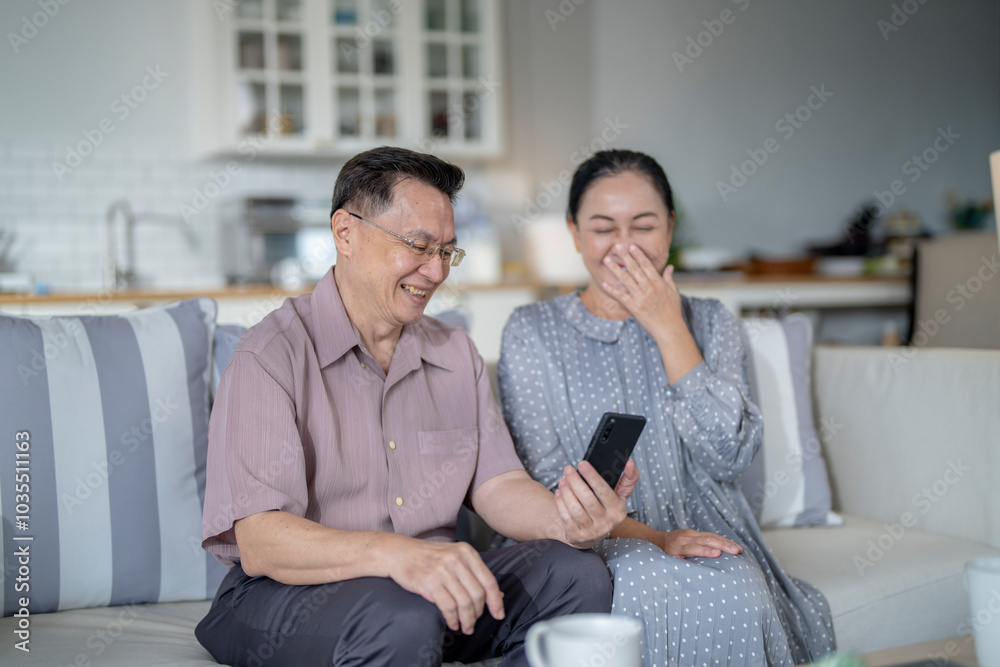 An elderly couple sits together in their cozy living room, smiling and enjoying a moment as they look at something on a smartphone. Their warmth and affection capture a peaceful home atmosphere. An elderly couple sits together in their cozy living room, smiling and enjoying a moment as they look at something on a smartphone. Their warmth and affection capture a peaceful home atmosphere.
