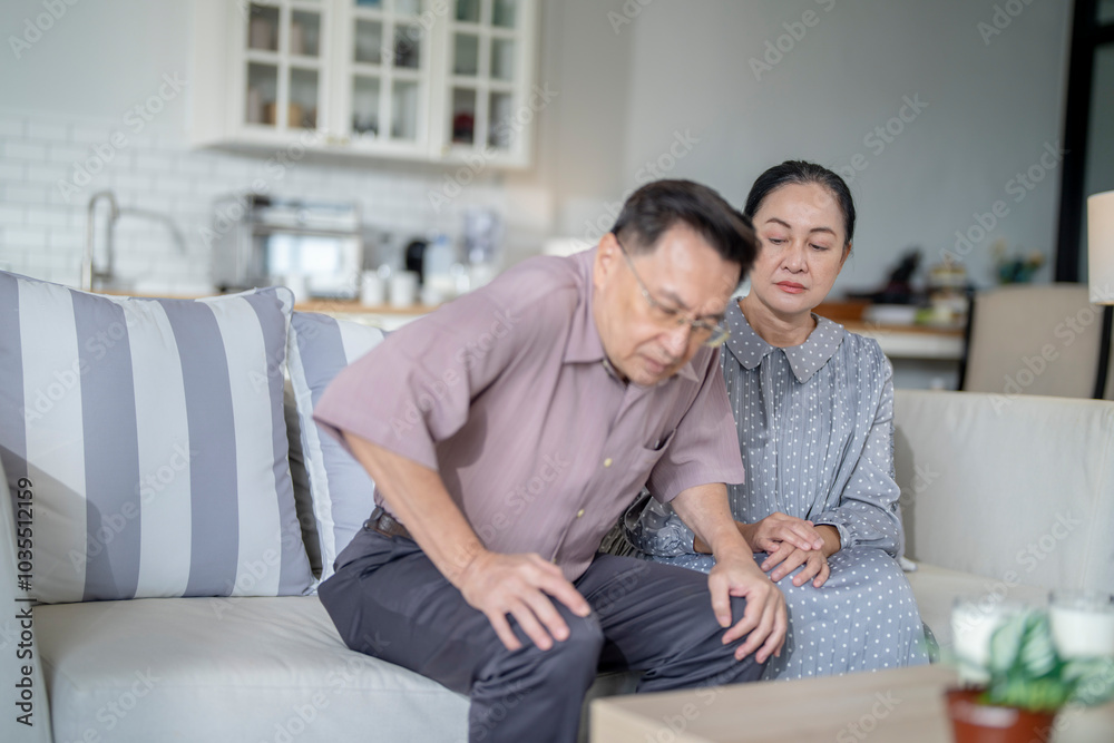 An elderly couple sits in their living room. The man holds his knee, appearing to be in discomfort, while his partner offers support. The image conveys care, concern, and the bond between the couple. An elderly couple sits in their living room. The man holds his knee, appearing to be in discomfort, while his partner offers support. The image conveys care, concern, and the bond between the couple.