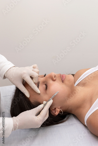 young woman on a table in a beauty center performing a beauty treatment for facial skin with a scalpel using the dermaplaning technique