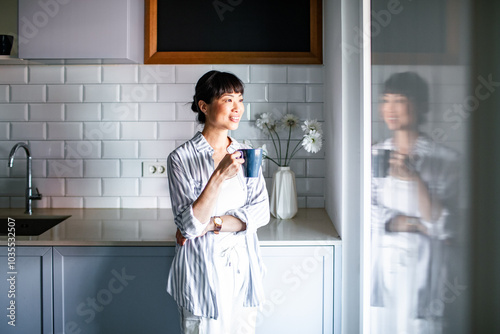 Asian woman drinking coffee in modern kitchen by window photo