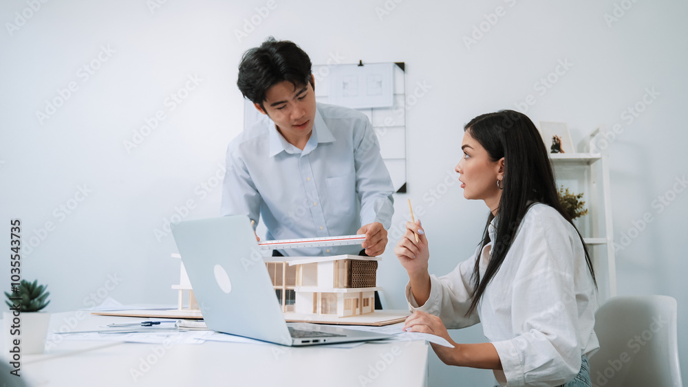 Professional male asian architect using ruler to measure house model length while young beautiful caucasian colleague using laptop to analyzed data on meeting table with house model. Immaculate. Professional male asian architect using ruler to measure house model length while young beautiful caucasian colleague using laptop to analyzed data on meeting table with house model. Immaculate.