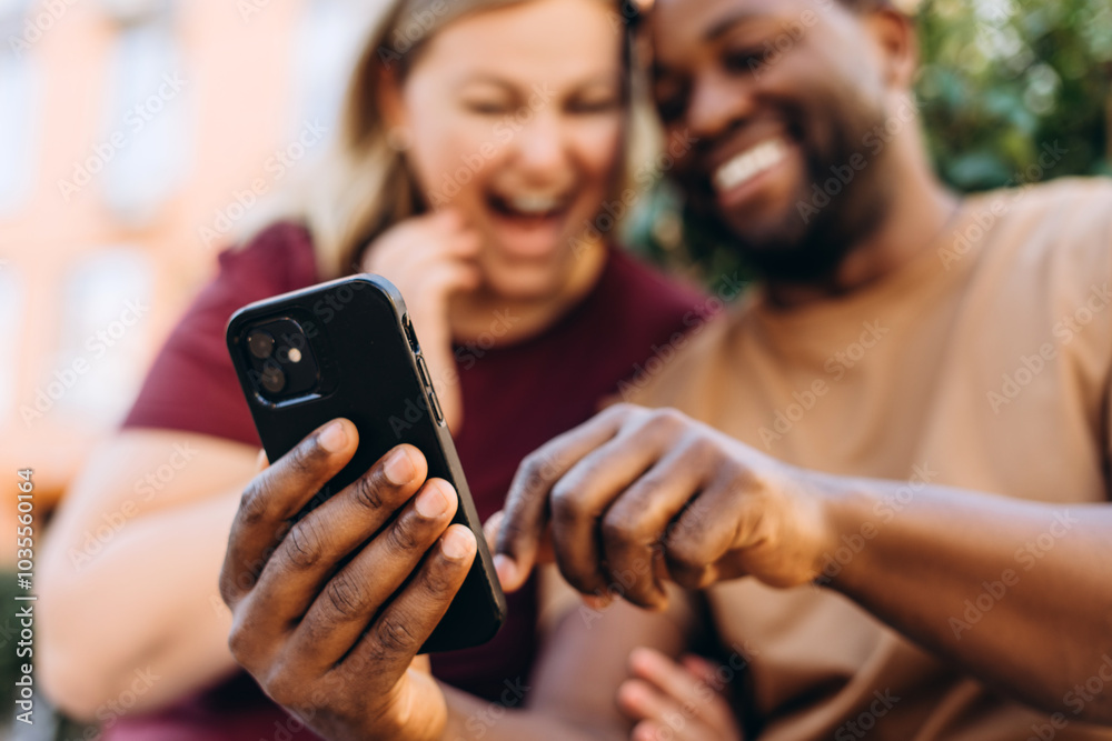 Fototapeta premium Happy multiethnic couple looking at smartphone screen sitting outside