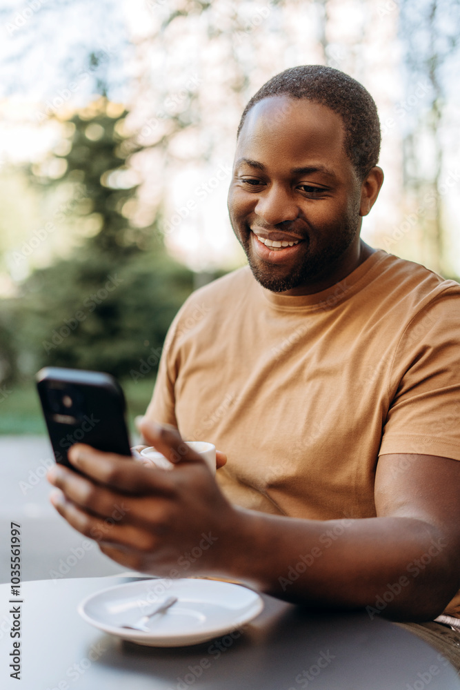 Fototapeta premium Happy African American man drinking coffee using smartphone at cafe outdoors