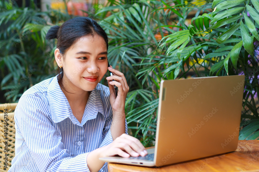 happy asian woman enjoy working on laptop and talking on mobile phone while sitting in outdoor cafe with green plant interior happy asian woman enjoy working on laptop and talking on mobile phone while sitting in outdoor cafe with green plant interior