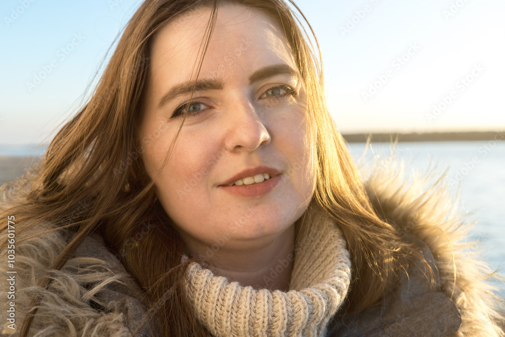 Close-up of a smiling woman in a winter jacket, with the soft glow of a sunset on a beach. The light highlights her relaxed expression, with a calm sea and sandy shore in the background Close-up of a smiling woman in a winter jacket, with the soft glow of a sunset on a beach. The light highlights her relaxed expression, with a calm sea and sandy shore in the background