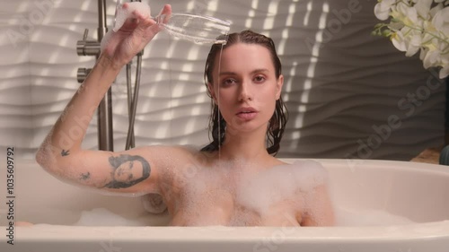 Relaxing sexy sensual naked Caucasian drunk woman female girl lady in spa bathing washing bath foam at home holding glass of drink pouring champagne pour alcohol in bathtub addiction looking at camera