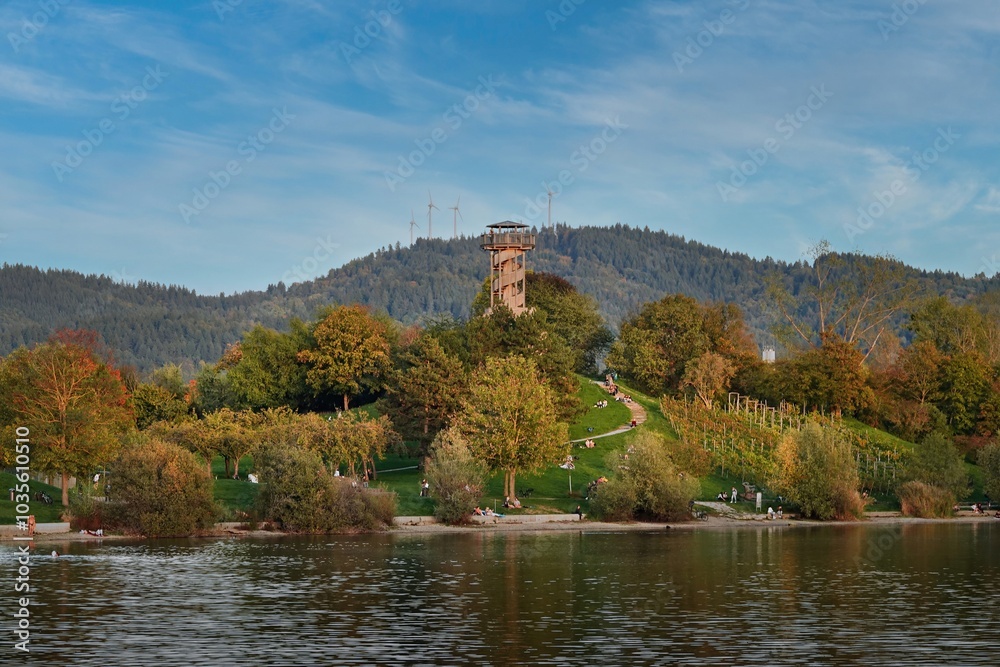 The last warm days of autumn. People enjoy the warmth on a sunlit hill at Freiburg’s Seepark, surrounded by autumn colors. Wind turbines in the background on the mountain