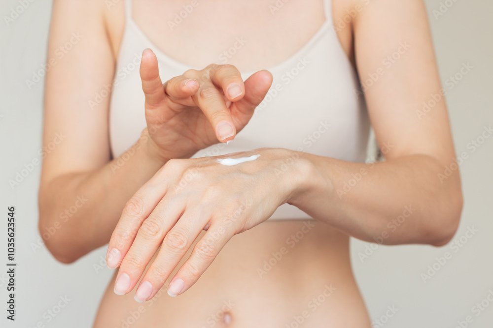 Fototapeta premium Young woman in beige underwear gently applying a white moisturizing and nourishing hand cream to the skin of her hands. Isolated on a beige background